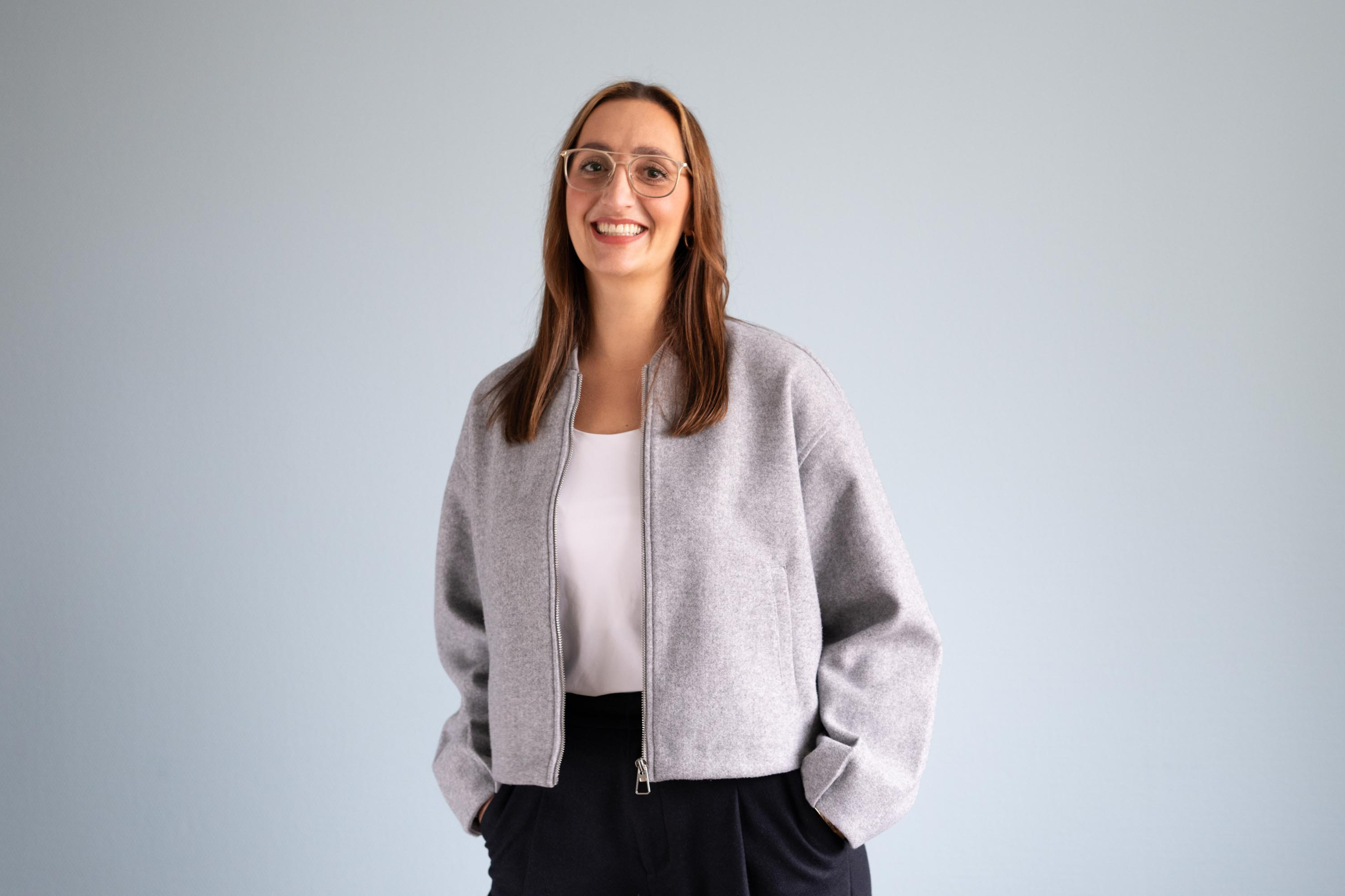 Eine junge Frau in grauer Jacke und mit Brille steht vor einer Wand und lächelt.