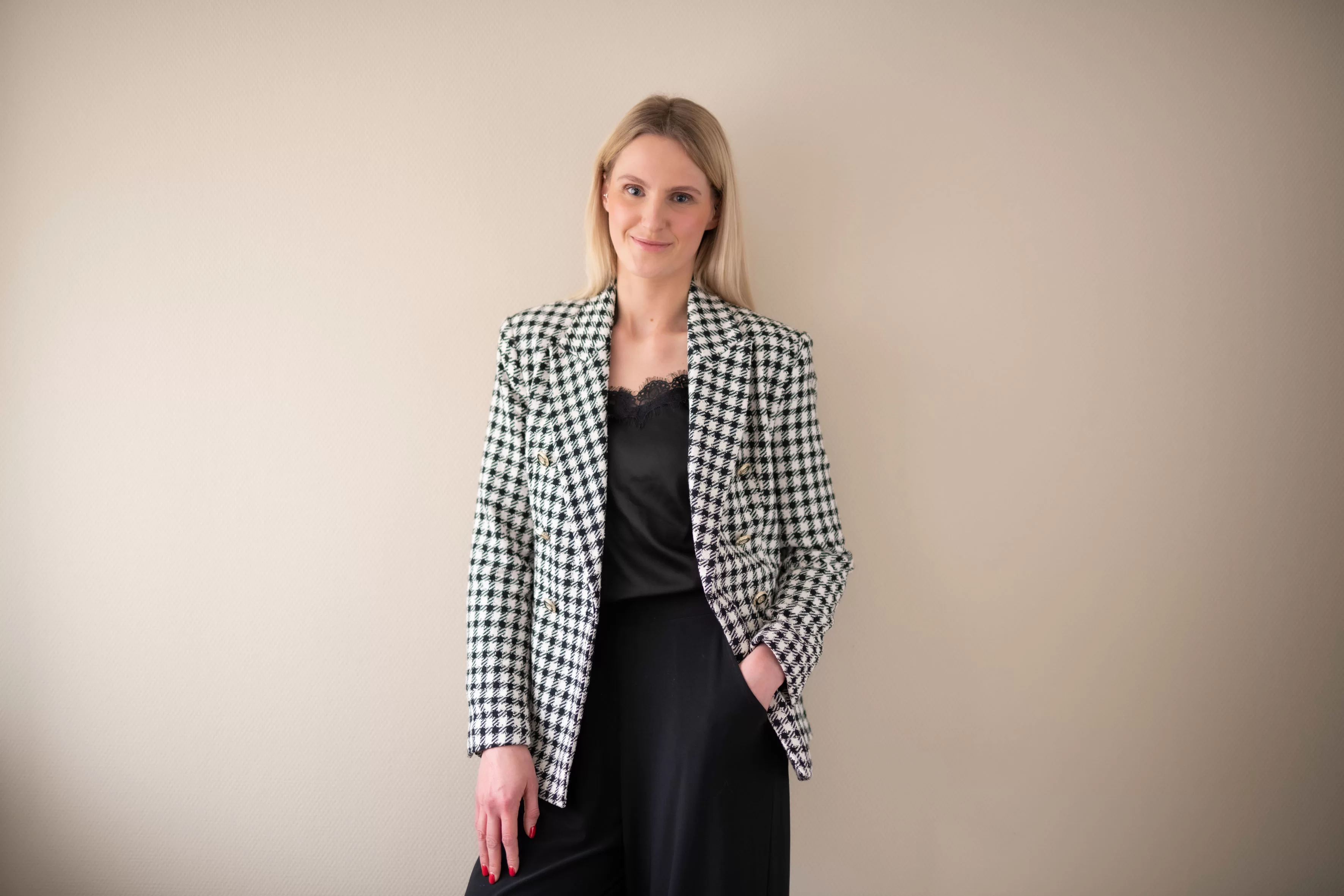 Eine blonde Frau in kariertem Blazer lehnt an der Wand und lächelt. 